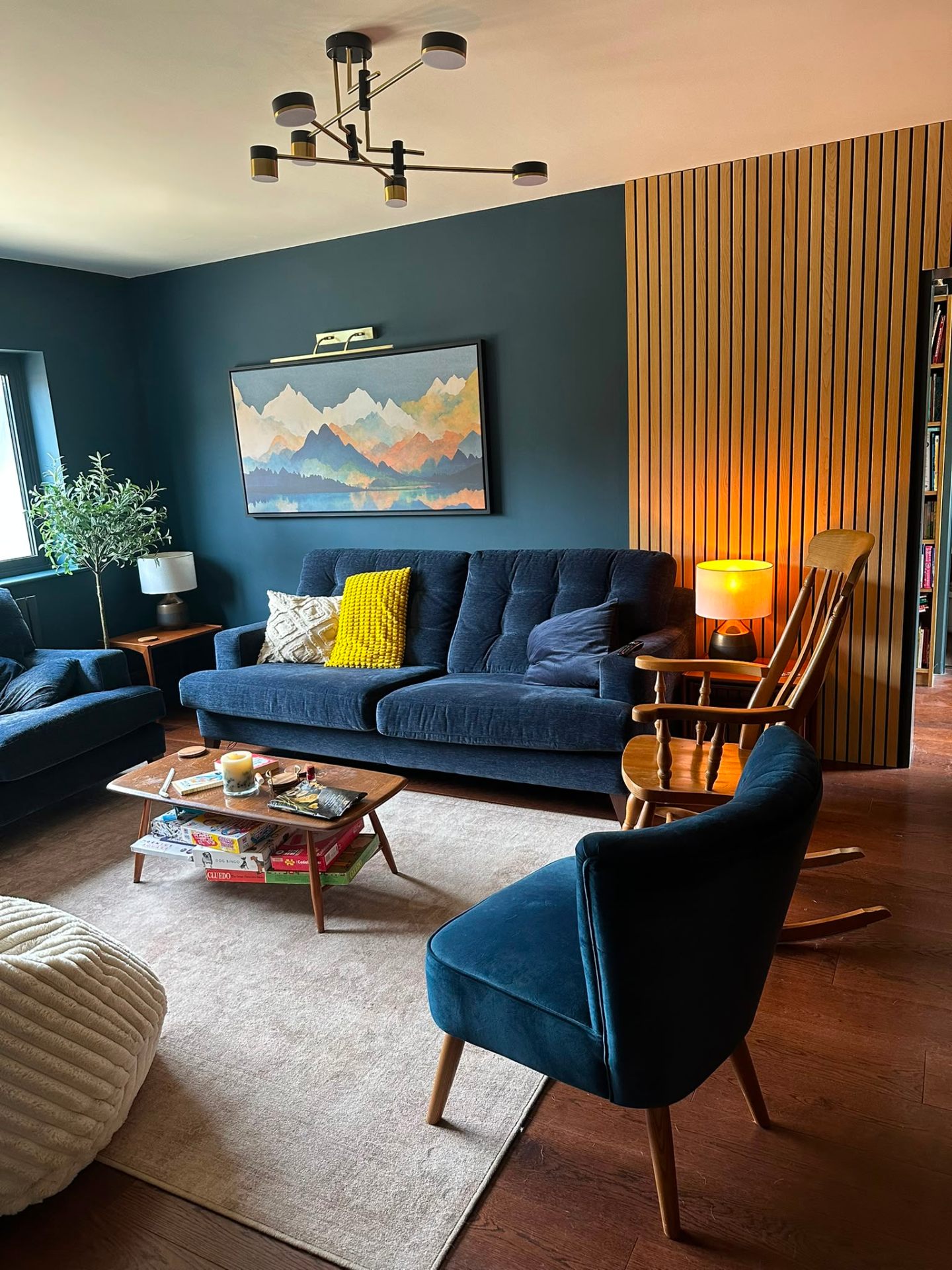 Sage-green snug with black-and-brass ceiling pendant, brass picture light and warm LED accent behind oak slat wall — living room lighting design by D.P.L Electrical Services